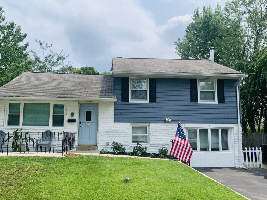A split-level house with blue and white exterior, a front lawn, and an American flag near the driveway.
