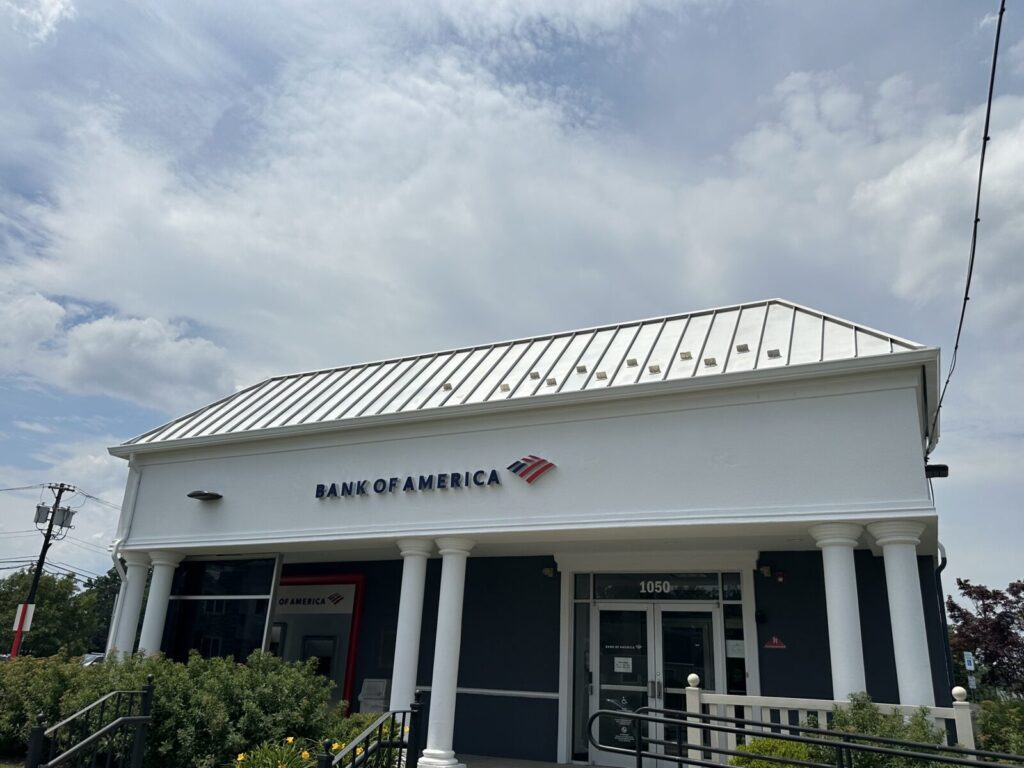 A Bank of America branch building with a white roof, columns, and clear signage under a partly cloudy sky.