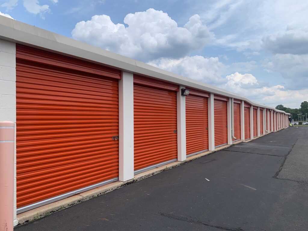 Row of orange storage unit doors at a storage facility, with a paved driveway and cloudy sky overhead.
