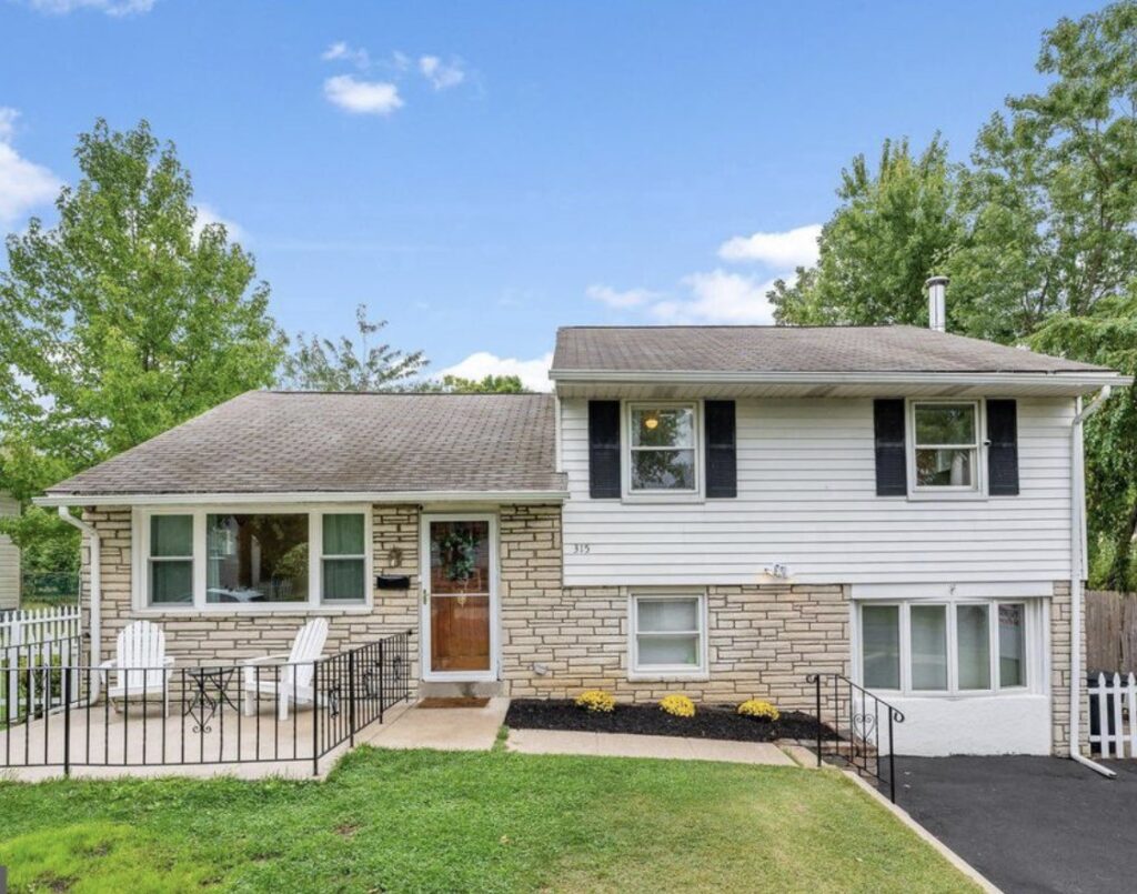 Split-level house with stone and siding exterior, black shutters, front porch, and a paved driveway, surrounded by trees.