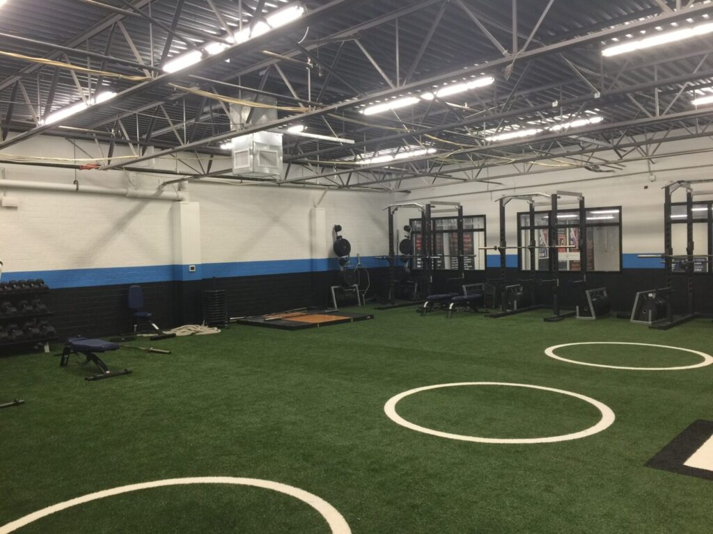 Indoor gym with artificial turf flooring, weightlifting racks, benches, and free weights against the walls.