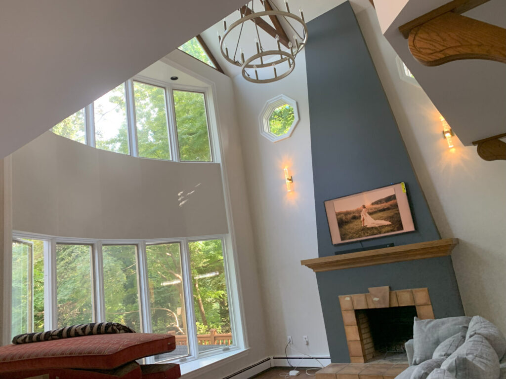 Living room with large curved windows, high ceiling, fireplace, modern chandelier, and a TV above the mantel.