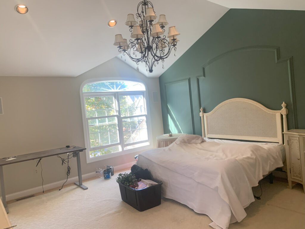 A bedroom with a covered bed, green accent wall, chandelier, large window, and miscellaneous items on the floor.