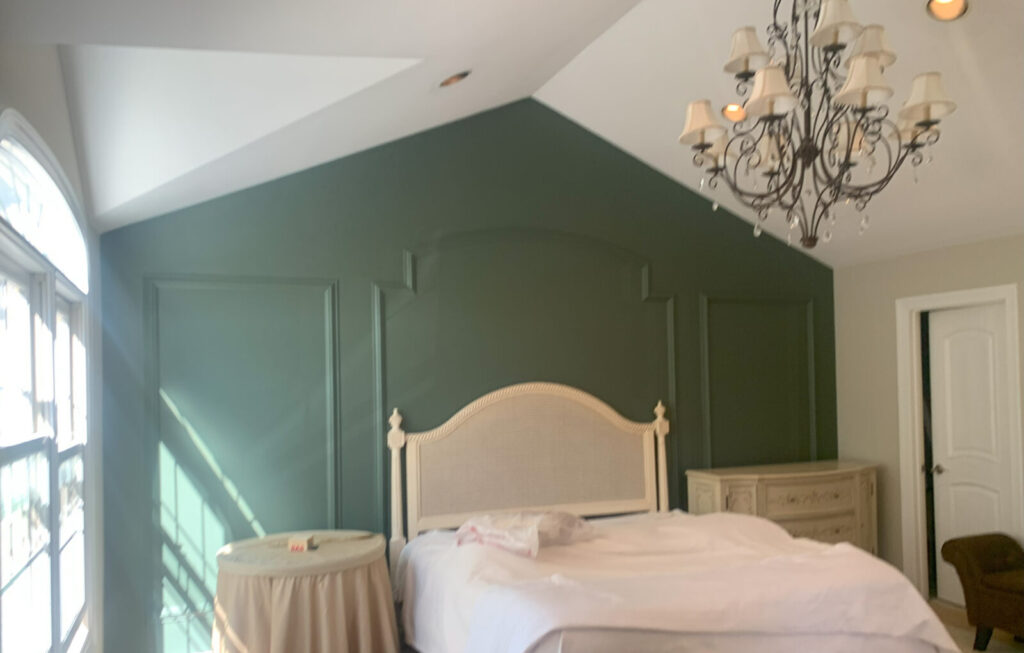 Bedroom with a green accent wall, chandelier, bed, round bedside table, and dresser under a vaulted ceiling.
