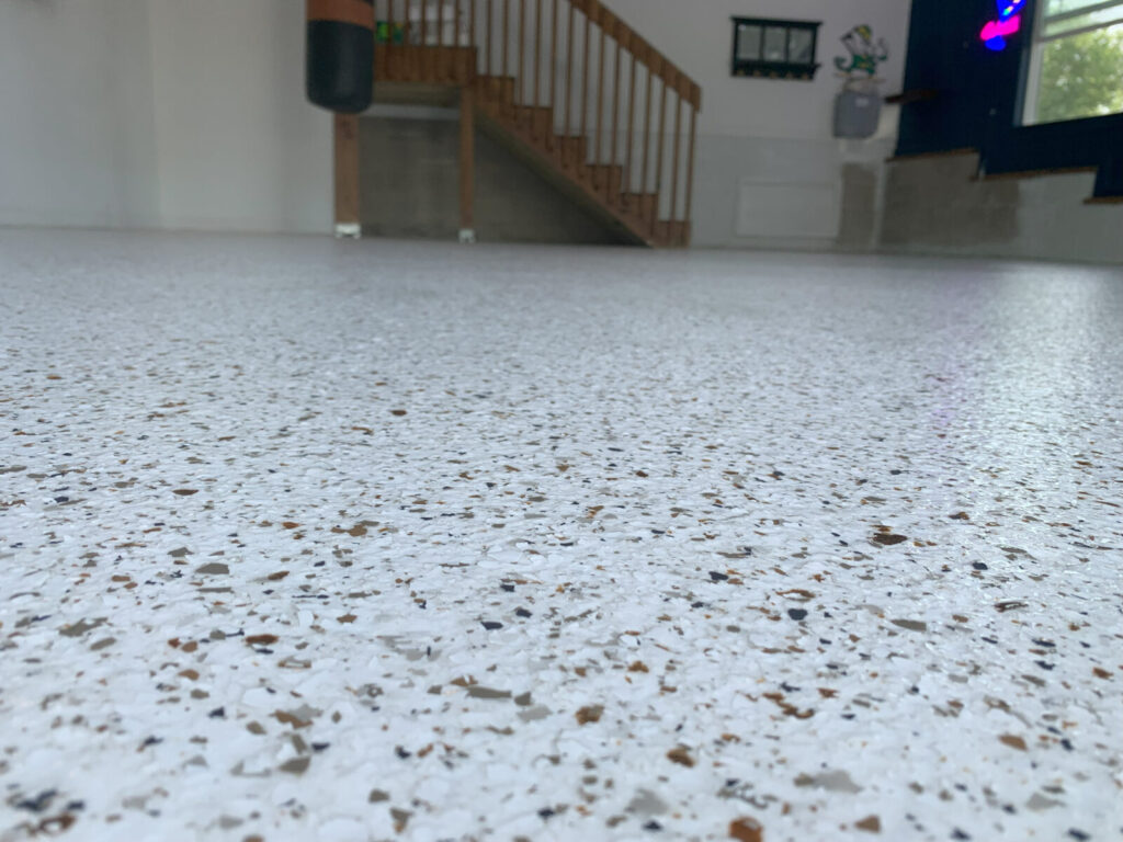 Close-up view of a speckled epoxy garage floor with stairs, railing, and gym equipment in the background.