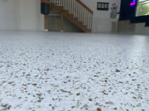 Close-up view of a speckled epoxy garage floor with stairs, railing, and gym equipment in the background.