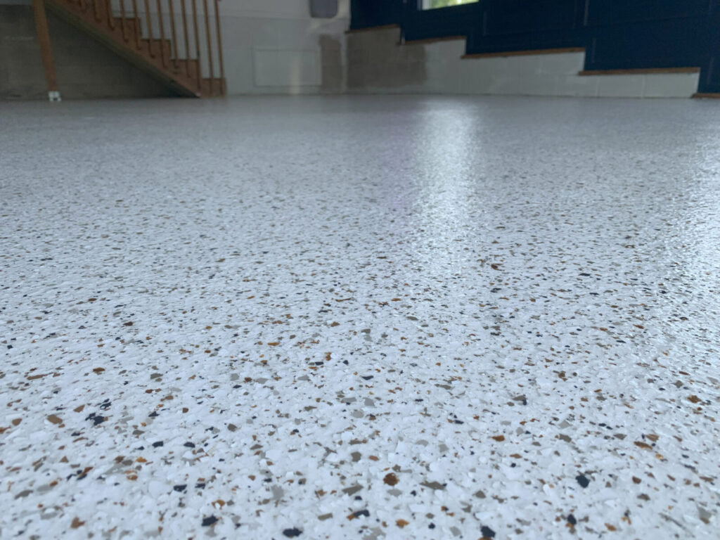 Close-up view of a speckled epoxy-coated floor in a room with stairs and blue and white walls in the background.